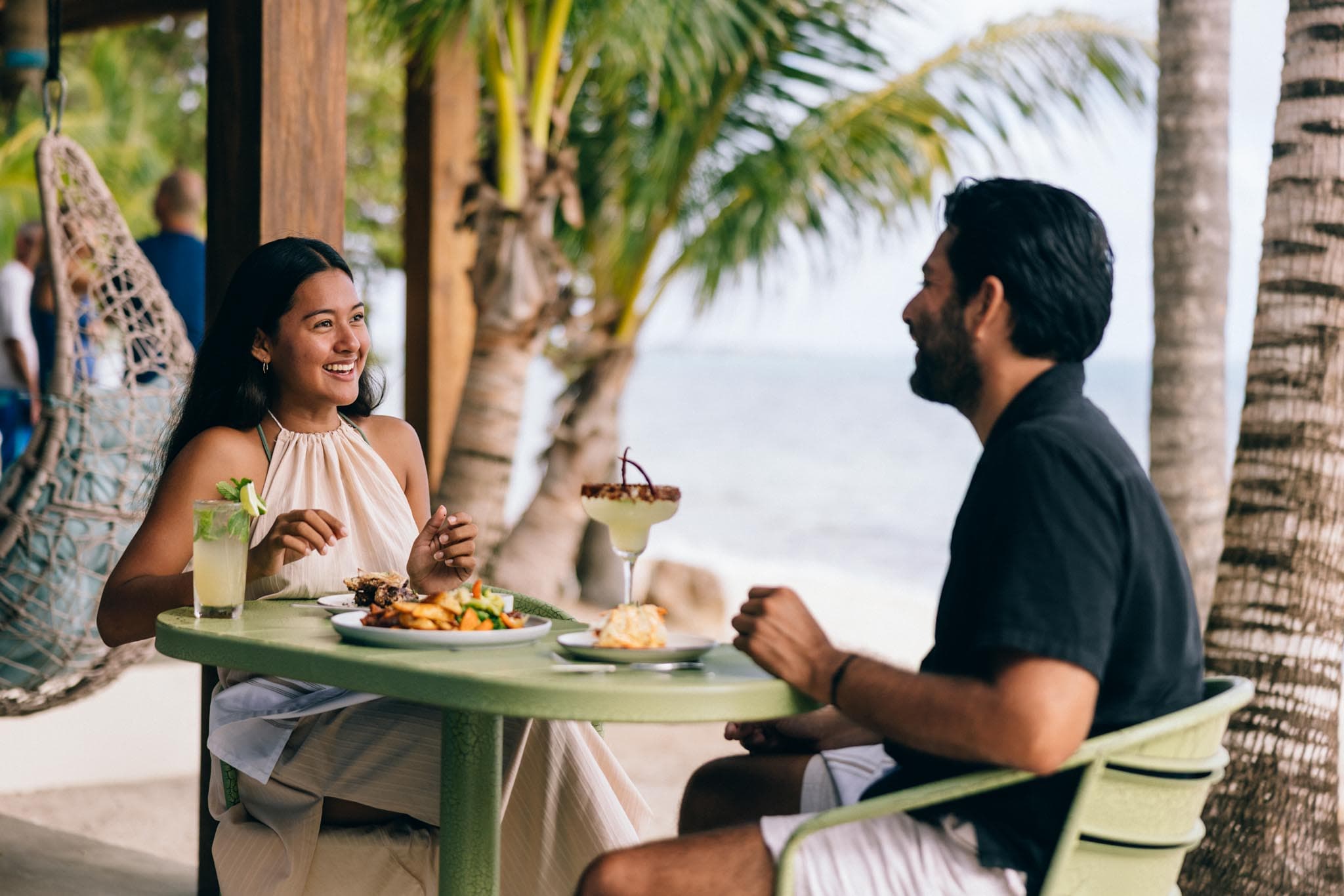 A woman and a man enjoy a meal at a beachside restaurant, smiling and engaged in conversation.